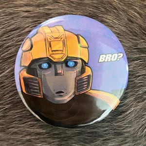 May include: Round button with a cartoon illustration of a robot's face, featuring yellow and gray colors with blue eyes. The button has the word "BRO?" in white text on a blue background.