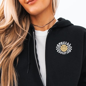 May include: Black zip-up hoodie with a white t-shirt underneath. The hoodie features a circular design on the upper left chest with the words "SPECIAL EDUCATION" around a smiley face on a checkered background. The model is wearing light blue jeans.