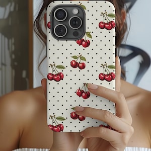 May include: A white phone case with a black polka dot pattern and a red cherry design. The case has a camera lens cutout and a cutout for the phone's buttons.
