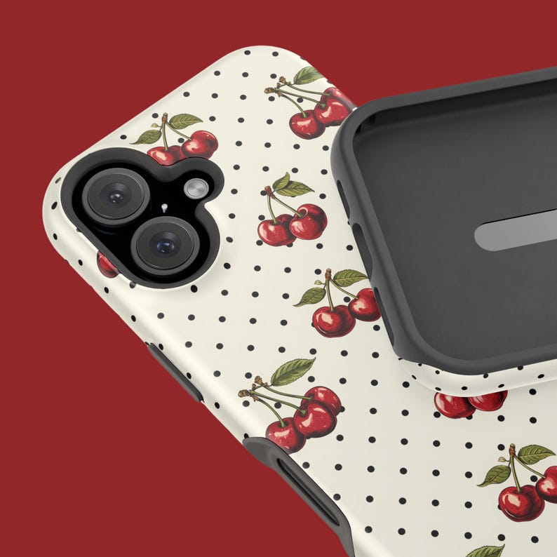 May include: A white phone case with a pattern of red cherries and black polka dots. The case has a raised camera bump with two lenses.