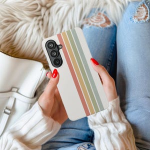 May include: A white phone case with vertical stripes in muted colors of red, yellow, green, and blue. The phone case is held in a person's hands, with red nail polish visible. The phone has a triple-lens camera.