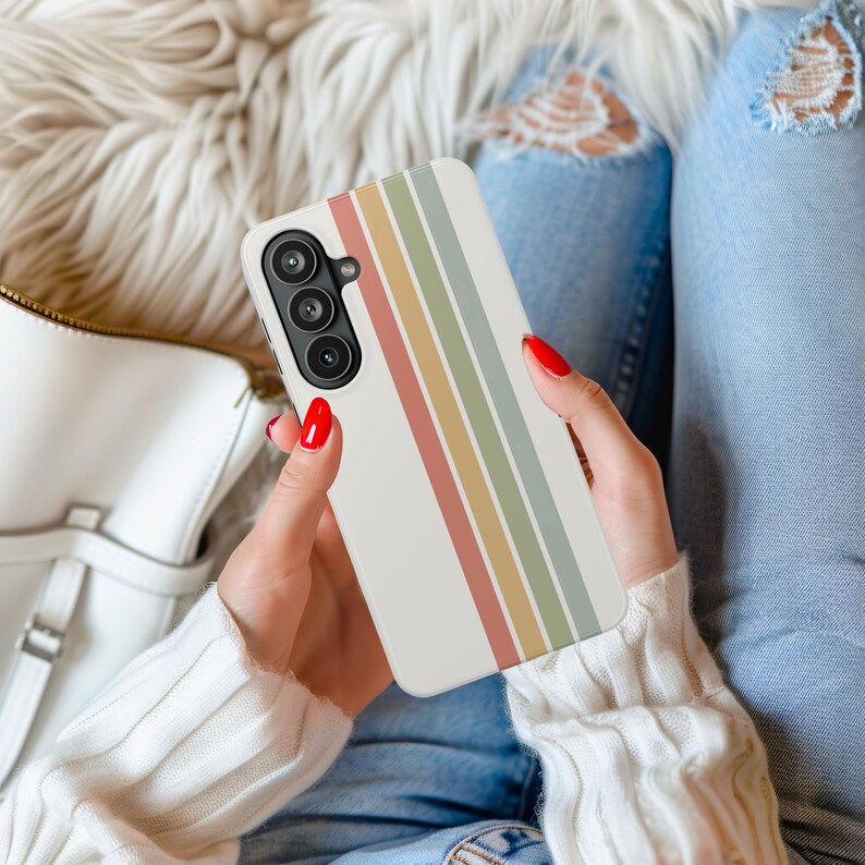 May include: A white phone case with vertical stripes in muted colors of red, yellow, green, and blue. The phone case is held in a person's hands, with red nail polish visible. The phone has a triple-lens camera.