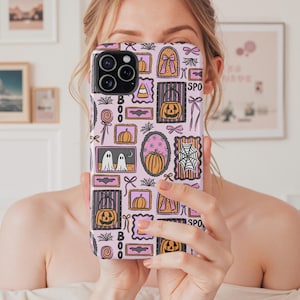May include: A pink phone case with a repeating Halloween-themed pattern. The design includes pumpkins, ghosts, spiderwebs, bows, and the word "BOO" in black. The case has a glossy finish and a camera cutout.