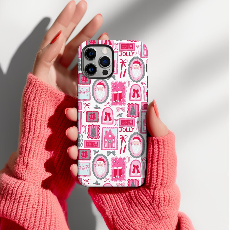 May include: A white phone case with a repeating pattern of pink Christmas-themed illustrations, including Santa Claus, bows, and houses. The word "JOLLY" is printed on the case. The case is held by a person wearing a pink sweater.