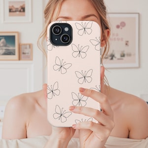 May include: A phone case with a light pink background and a repeating pattern of black line-drawn butterflies. The case has a camera cutout and is held up in front of a person's face.