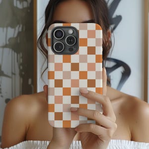 May include: A smartphone case featuring a repeating checkered pattern in shades of burnt orange, light peach, and cream. The design covers the entire back of the case, which has a cutout for a triple-lens camera system located in the upper left corner.
