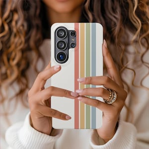 May include: A white smartphone case with a vertical striped design in shades of red, orange, yellow, green, and blue. The case is held in front of a person, showcasing the phone's camera array.