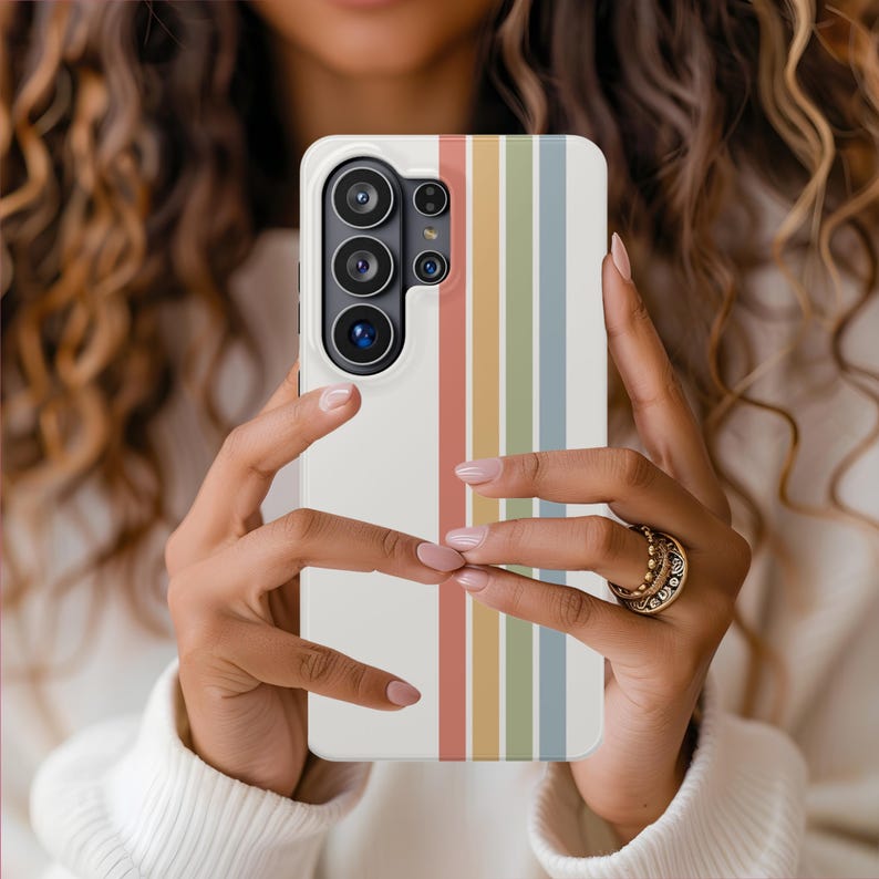 May include: A white smartphone case with a vertical striped design in shades of red, orange, yellow, green, and blue. The case is held in front of a person, showcasing the phone's camera array.