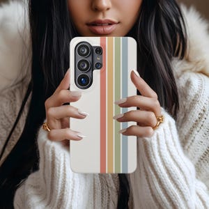 May include: A white smartphone case with vertical stripes in pastel colors: red, yellow, green, and blue. The case has a camera module with multiple lenses. The phone case is held by a person wearing a white sweater.