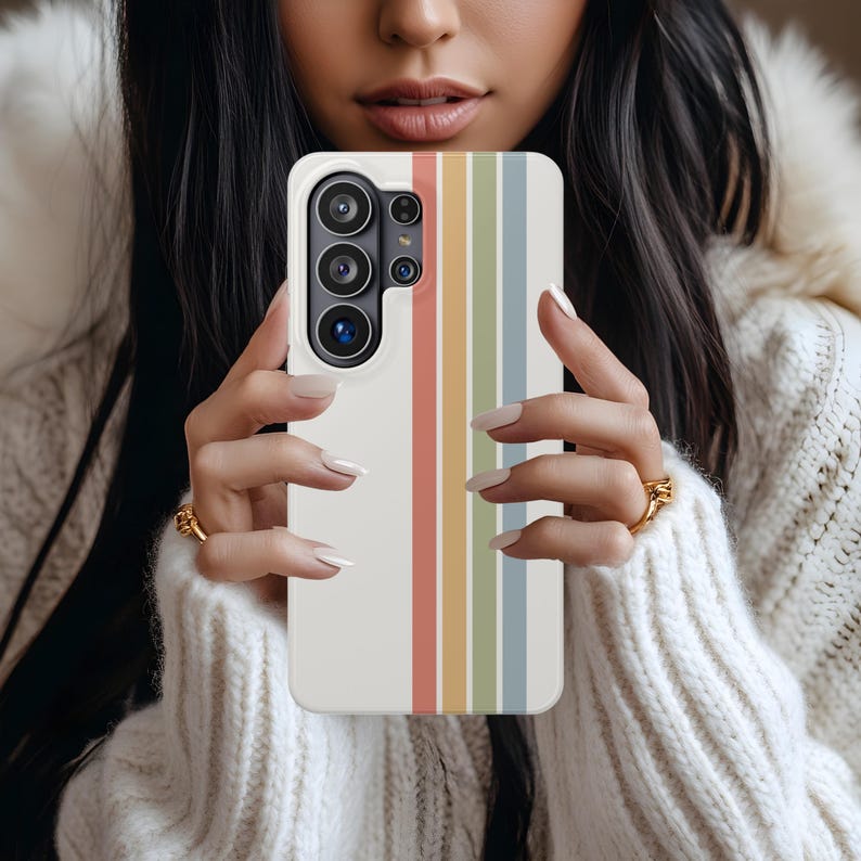 May include: A white smartphone case with vertical stripes in pastel colors: red, yellow, green, and blue. The case has a camera module with multiple lenses. The phone case is held by a person wearing a white sweater.