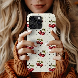 May include: A white phone case with a black polka dot pattern and a repeating image of red cherries. The case has a camera lens cutout with three black circles.