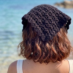 May include: A black triangular headscarf featuring a textured, repeating crochet pattern. The accessory is crafted from yarn and displays a distinct, open-work stitch design that creates a geometric, grid-like appearance across the fabric.