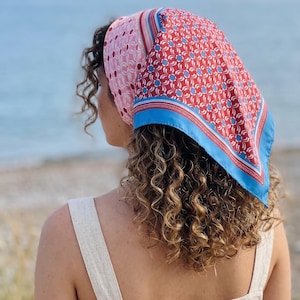 May include: A square headscarf featuring a vibrant pattern of red, white, and blue floral motifs, accented by a light pink section with a repeating circular design. The scarf is finished with a solid light blue border and thin red stripes.
