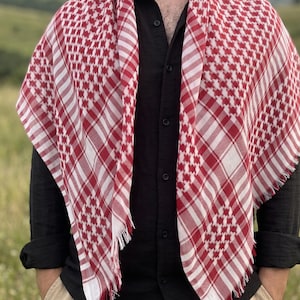 Może przedstawiać: Czerwono-biała szachownica keffiyeh narzucona na czarną koszulę. Szal ma postrzępioną krawędź i geometryczny wzór. Osoba ma na sobie czarną koszulę zapinaną na guziki i beżowe spodnie. Tło to rozmyta sceneria na zewnątrz.