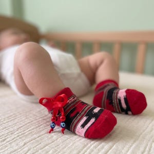 Könnte beinhalten: Ein Paar Socken mit rotem, schwarzem und rosafarbenem Muster, die am Knöchelbereich mit einer kleinen roten Schleife versehen sind. Zwei kleine blau-weiße Nazar-Perlen hängen an der roten Schleife und bilden ein dekoratives Detail am Socken.