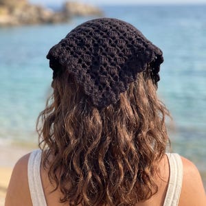 May include: A black triangular headscarf featuring a textured crochet pattern with a scalloped edge. The accessory is crafted from yarn and designed to be worn over the hair, covering the crown with a repeating geometric stitch construction.