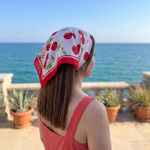 May include: A white headscarf with a red border and a pattern of red cherries and green leaves. The headscarf is tied around the head. The person is wearing a red dress and looking out at the ocean.