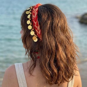 Puede incluir: Una diadema trenzada roja y dorada con monedas de oro colgantes. La diadema la lleva una persona con cabello castaño largo y ondulado. El fondo muestra el mar.