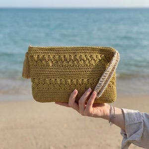 May include: A mustard-colored crochet clutch with a textured pattern and a matching tassel. The clutch has a cream-colored wrist strap and is held up against a backdrop of the ocean.