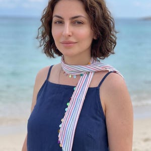 May include: A dark blue sleeveless top with thin straps, paired with a long, narrow scarf featuring thin vertical stripes in red, green, and blue. The scarf is adorned with small, colourful circular metallic discs along its edges.