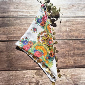 Op de afbeelding: Een witte stoffen sjaal met een kleurrijk paisley-patroon en gouden kwastjes. De sjaal is driehoekig gevouwen en ligt op een houten oppervlak.
