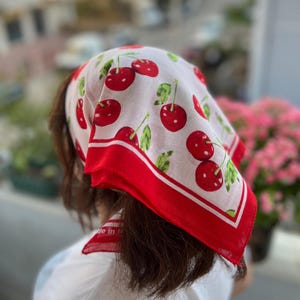 May include: White headscarf with a red cherry pattern and a red border. The scarf is made of a lightweight fabric and is tied around the head. The scarf has the words "Made in" printed on it.