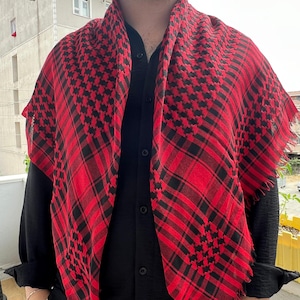 May include: A red and black chequered scarf draped over a black button-down shirt. The scarf has a fringed edge and a geometric pattern. The outfit is completed with beige trousers.