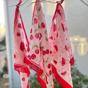 May include: Three light pink bandanas with red borders and fruit patterns, including strawberries, cherries, and watermelon slices, are tied to a white branch. The bandanas are hanging in front of a blurred outdoor background.