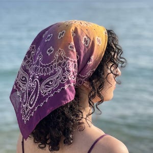 Puede incluir: Un pañuelo cuadrado con un degradado de color que va del morado intenso al amarillo brillante. La tela está decorada con un patrón de cachemira blanco tradicional, que incluye intrincados remolinos y motivos florales.