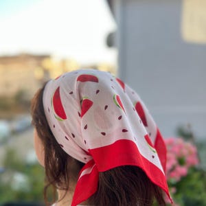 May include: A white bandana with a red watermelon slice pattern and a red border. The bandana is tied around the head, with the design visible. The bandana is made of a lightweight fabric.