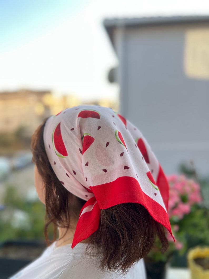 May include: A white bandana with a red watermelon slice pattern and a red border. The bandana is tied around the head, with the design visible. The bandana is made of a lightweight fabric.