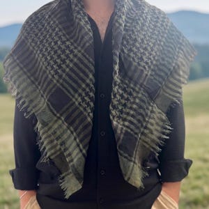 Peut inclure: Écharpe à carreaux noirs et vert olive avec des bords effilochés. L'écharpe est drapée sur une chemise noire boutonnée. L'arrière-plan présente un champ vert et des montagnes.