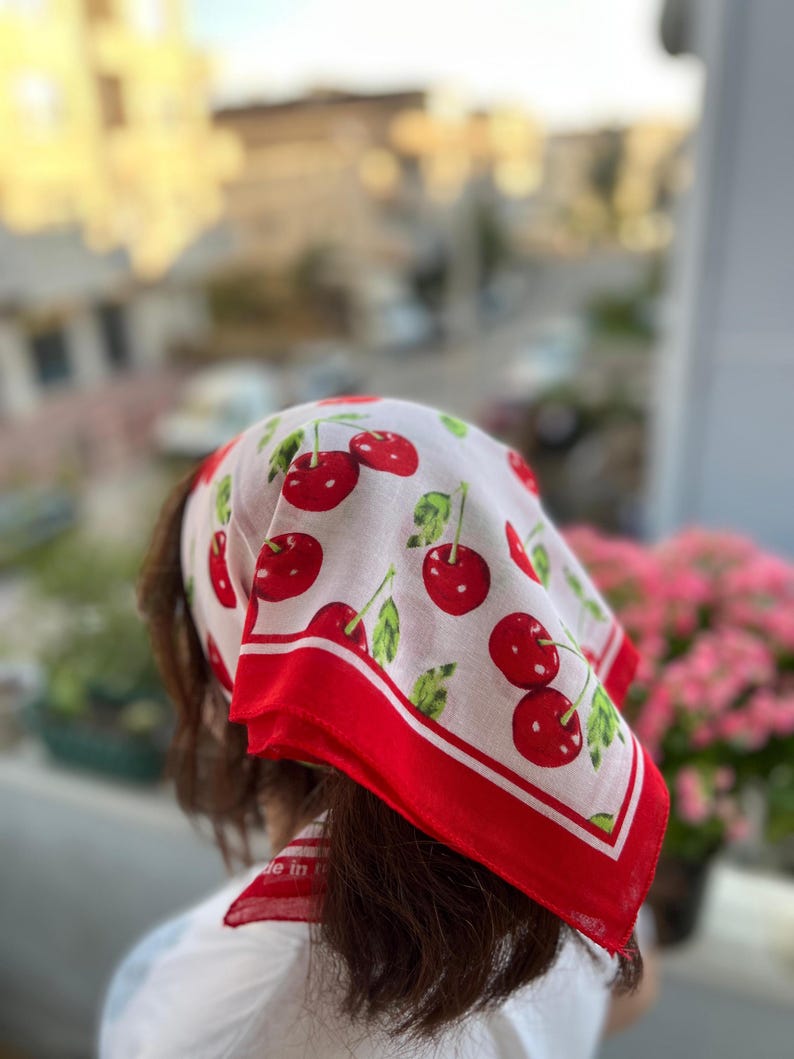 May include: White headscarf with a red cherry pattern and a red border. The scarf is made of a lightweight fabric and is tied around the head. The scarf has the words "Made in" printed on it.
