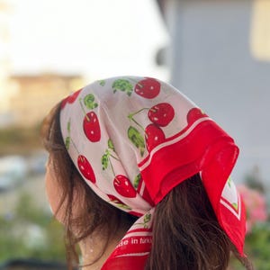 May include: A white headscarf with a red border and a pattern of red cherries and green leaves. The scarf is tied around the head, with the text "de in t&uuml;rki" visible on the corner.