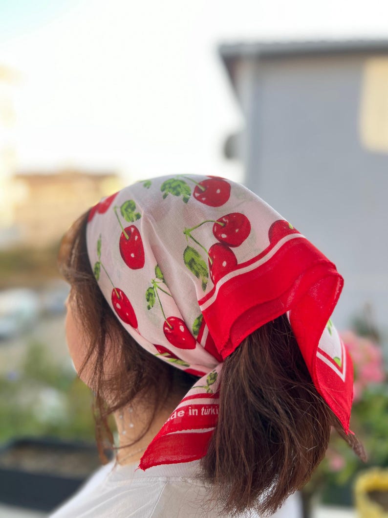 May include: A white headscarf with a red border and a pattern of red cherries and green leaves. The scarf is tied around the head, with the text "de in t&uuml;rki" visible on the corner.