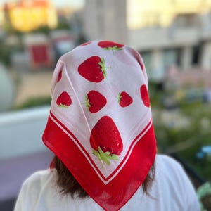 May include: A square, white headscarf with a repeating pattern of large red strawberries and green leaves. The scarf has a red border with a white stripe. The scarf is worn on the head.
