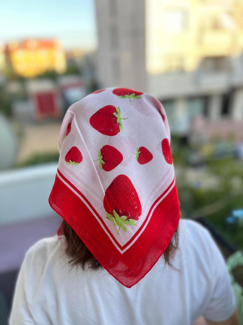 May include: A square, white headscarf with a repeating pattern of large red strawberries and green leaves. The scarf has a red border with a white stripe. The scarf is worn on the head.