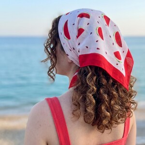 May include: A white headscarf with a red border and a pattern of red watermelon slices and black seeds. The scarf is tied at the back of the head. The person is wearing a red top.