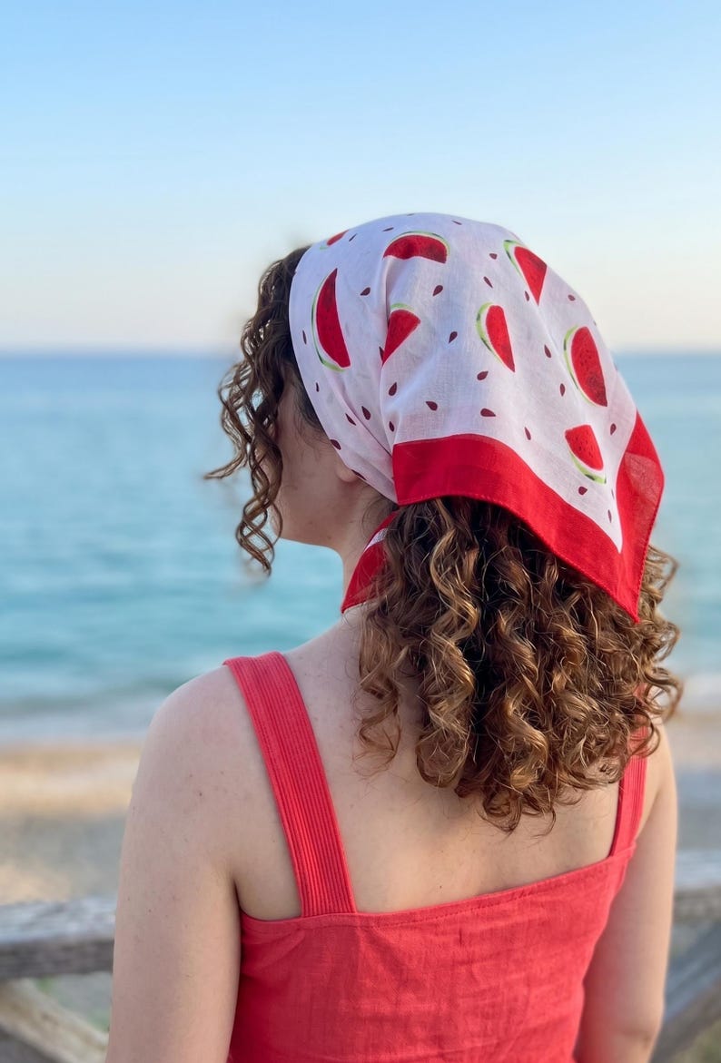 May include: A white headscarf with a red border and a pattern of red watermelon slices and black seeds. The scarf is tied at the back of the head. The person is wearing a red top.