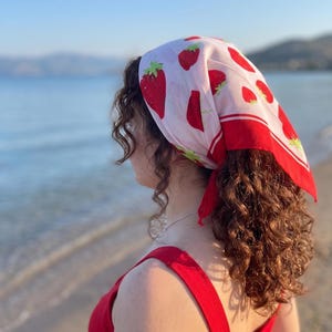 May include: A red and white strawberry print headscarf with a red border. The headscarf is tied at the back of the head. The person is wearing a red top. The background is a beach with the ocean and mountains.