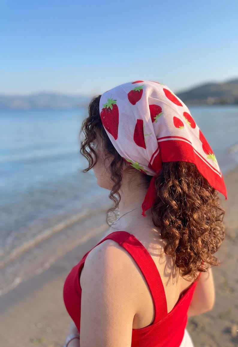 May include: A red and white strawberry print headscarf with a red border. The headscarf is tied at the back of the head. The person is wearing a red top. The background is a beach with the ocean and mountains.