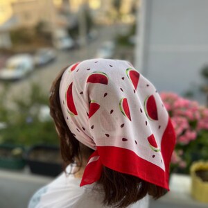May include: A white bandana with a red watermelon slice pattern and a red border. The bandana is tied around the head. The bandana is made of a lightweight fabric.