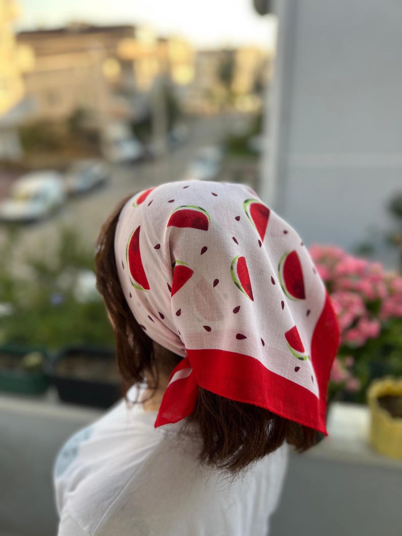 May include: A white bandana with a red watermelon slice pattern and a red border. The bandana is tied around the head. The bandana is made of a lightweight fabric.