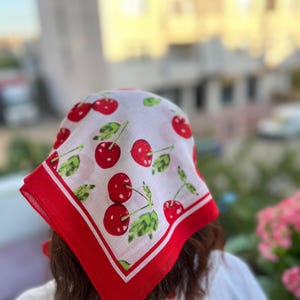 May include: A white bandana with a red border and a pattern of red cherries with green leaves. The bandana is square-shaped and appears to be made of a lightweight fabric.