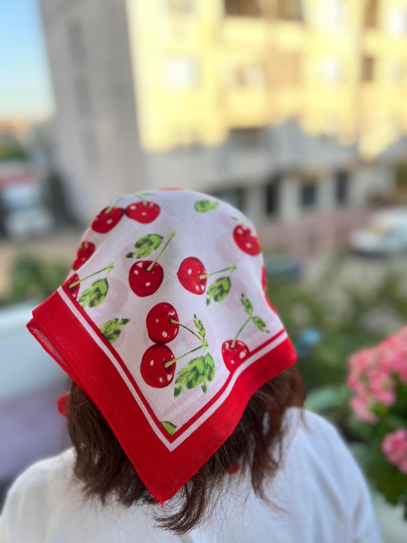 May include: A white bandana with a red border and a pattern of red cherries with green leaves. The bandana is square-shaped and appears to be made of a lightweight fabric.