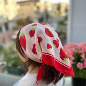 May include: A white headscarf with a repeating pattern of red strawberries and green leaves. The scarf has a red border and is tied at the back of the head. The scarf is worn by a person.