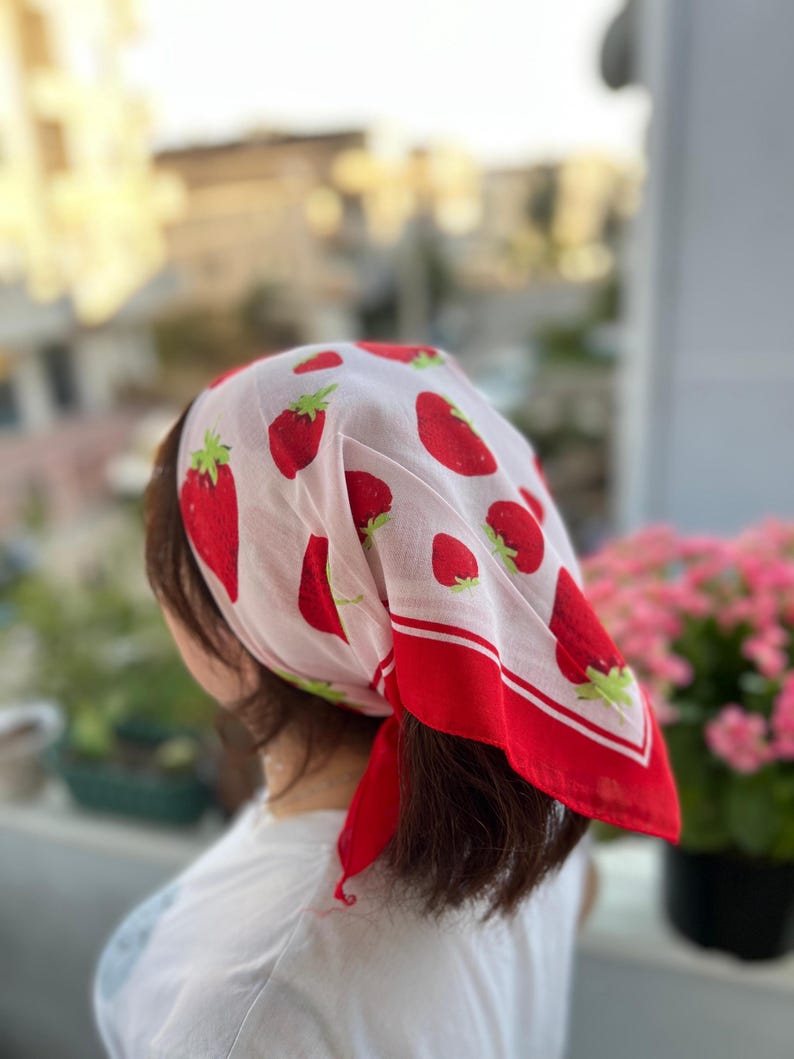 May include: A white headscarf with a repeating pattern of red strawberries and green leaves. The scarf has a red border and is tied at the back of the head. The scarf is worn by a person.