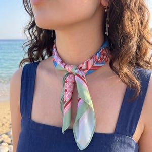 May include: A square neck scarf tied in a knot around the neck, featuring a multi-colored geometric and floral pattern in shades of sage green, light pink, red, and blue, worn with a dark blue sleeveless top with wide shoulder straps.