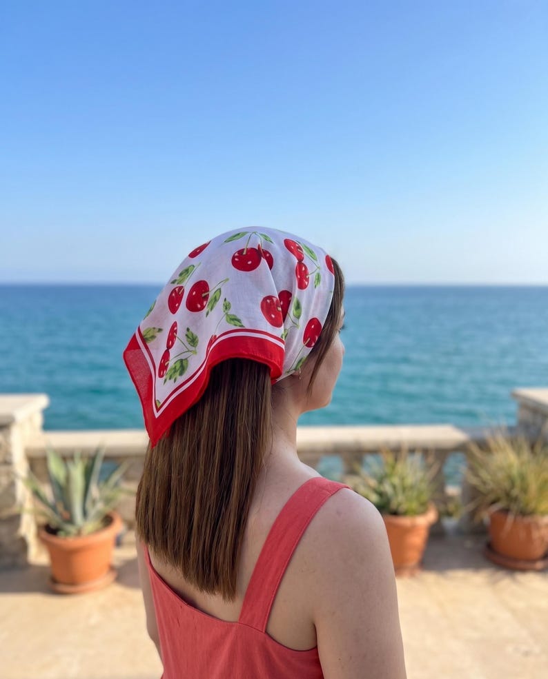 May include: A white headscarf with a red border and a pattern of red cherries and green leaves. The headscarf is tied around the head. The person is wearing a red dress and looking out at the ocean.