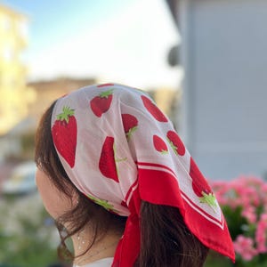 May include: A white headscarf with a red strawberry pattern and a red border. The strawberries are in various sizes and have green stems. The scarf is tied around the head.